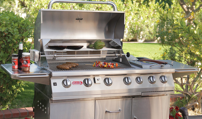 Stainless steel bull bbq grill cart grilling sausages, skewers, and eggplants outside with a green grass field background