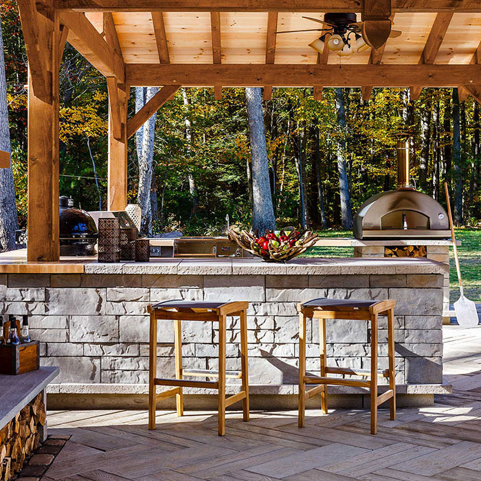 Close up of a grey colored tiled rock bar with wooden stools and designed in an outdoor kitchen including a grill, oven, and wooden roof with a forest in the background