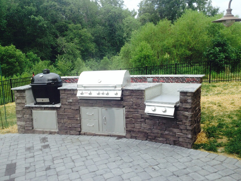 Outdoor kitchen with a Big Green Egg smoker grill next to a stainless steel grill and storage with a green tree forest background in Charlotte, North Carolina