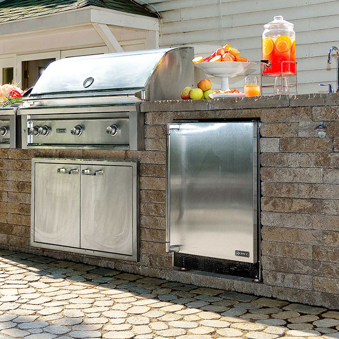 Close up of an outdoor grey colored tiled rock counter with a stainless steel grill and an adjacent stainless steel refrigerator with red and green apple water punch above the refrigerator