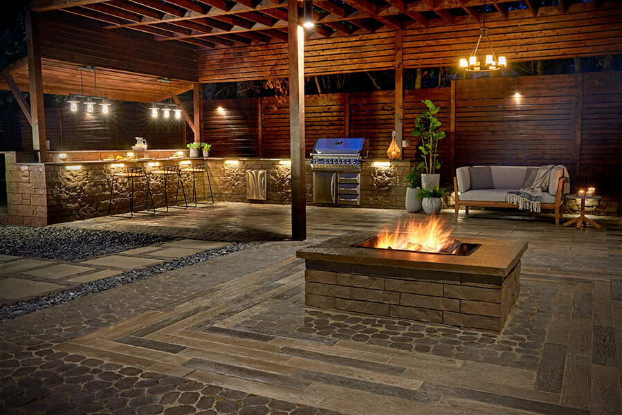 Night time capture of a stone tile fire pit surrounded by an outdoor kitchen with a grill and high top table adjacent to a grey outdoor couch