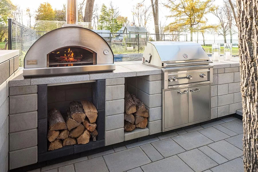 Close up of a stainless steel oven burning firewood next to a stainless steel grill designed in a stone tiled outdoor kitchen with log storage under the oven