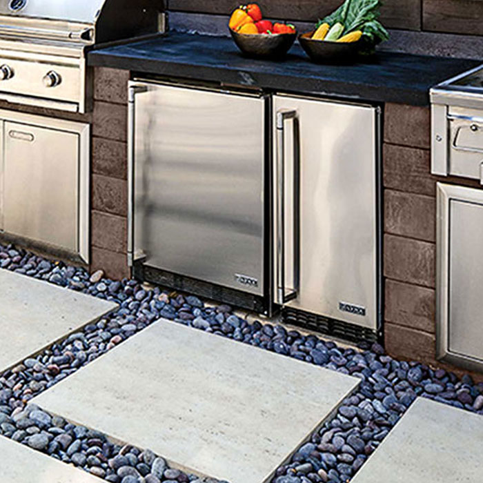 Close up of a shiny refrigerator designed in an outdoor kitchen placed in between a grill and a stove and in front of stone steps and pebbles