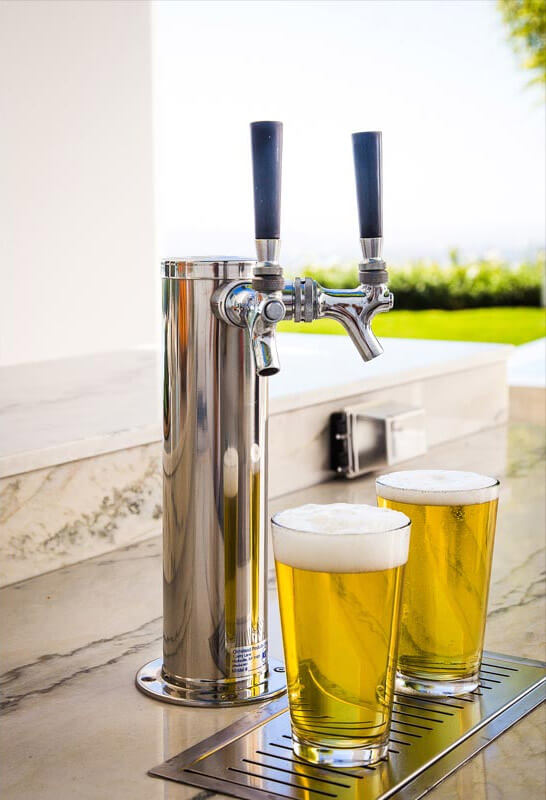 Close up of a double faucet stainless steel beer dispenser with two beer glasses filled with light beer and foam designed in an outdoor white marble kitchen design