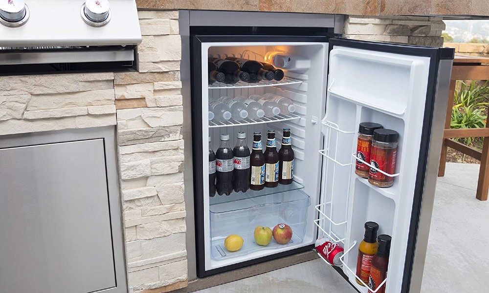Close up of an open stainless steel bull refrigerator in a white rock tile island with wine, water, soda, beer, apples, and hot sauce in different compartments