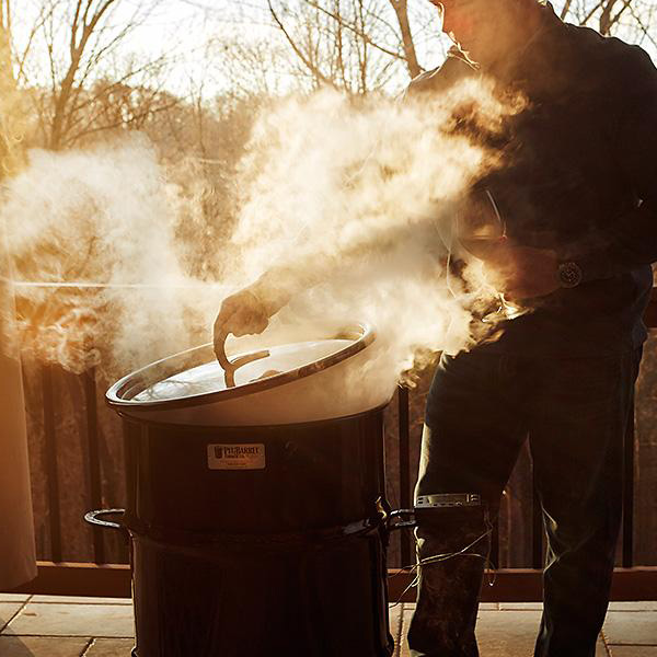 Close-up of a man opening a black painted metal 18.5 IN Classic Pit Barrel cooker that is steaming open