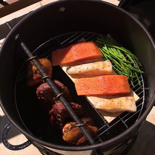Close-up of a black painted metal 18.5 IN Classic Pit Barrel cooker cooking four chickens on hooks and four salmon slices with asparagus