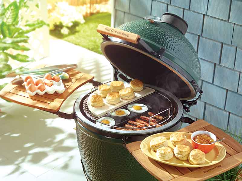 Close up capture of a Big Green Egg Cast Iron Plancha cooking three eggs sausages and heating biscuits with two side wooden tables