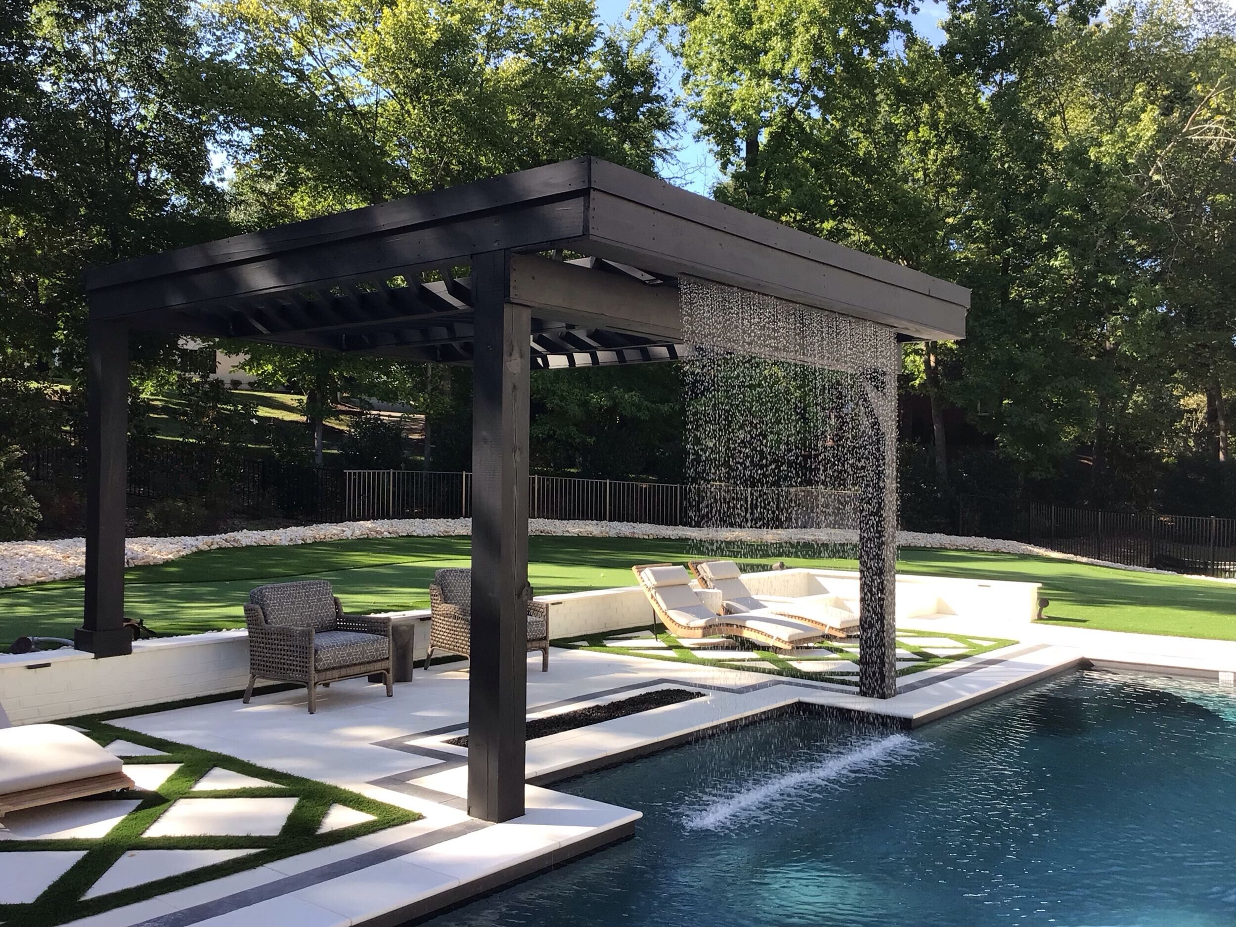 A stylish outdoor living space featuring a modern black pergola with a cascading water feature flowing into a luxurious pool, surrounded by lounge chairs, artificial turf, and elegant patio seating.