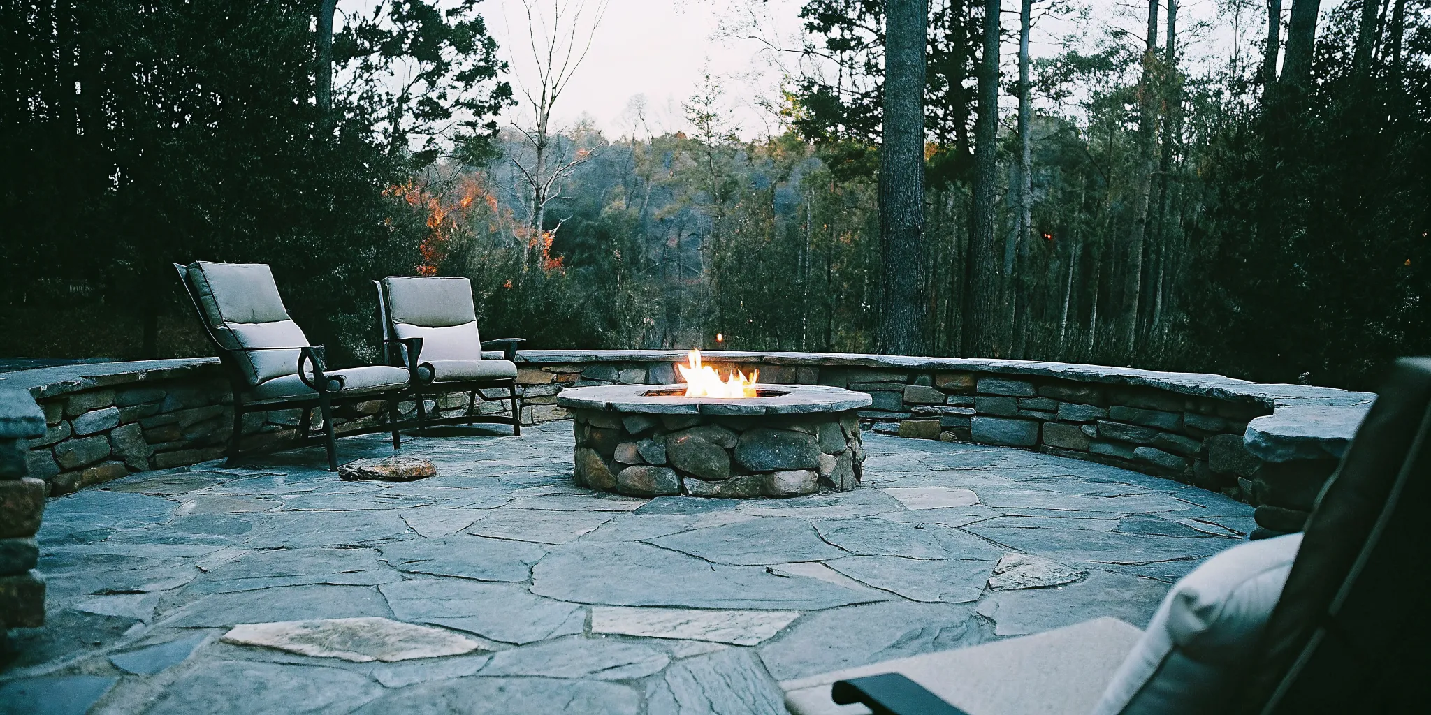 Custom stone outdoor fireplace and fire pit in a North Carolina backyard at dusk with warm glowing flames